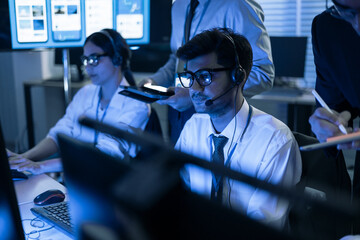 Professional call center operators work in a modern tech office. Team agent in headset monitors digital data while a supervisor provides technical guidance. Cybersecurity, network safety, and digital 