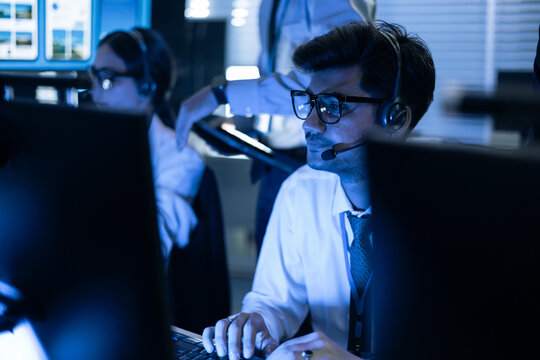 Focused male security operator with headset monitors data on screens in a high-tech control room. Professional team managing cybersecurity, network safety, and digital system operations daily