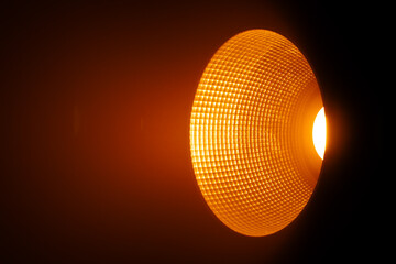 Close up of a studio spotlight emitting warm orange light with a textured reflector in a dark surrounding space. Lighting equipment backstage.