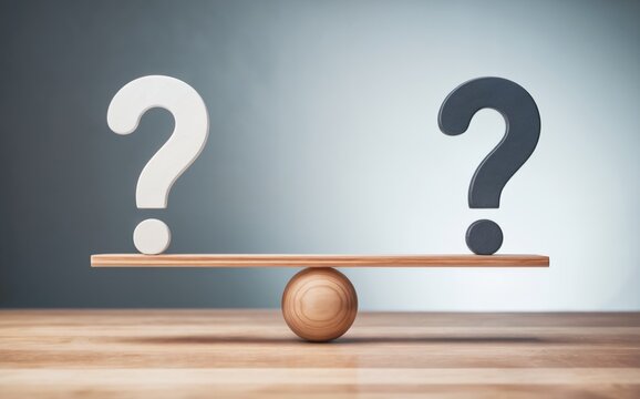 Two different colored question marks stand perfectly balanced on a simple wooden seesaw scale on a light wooden table against a softly lit grey studio background indicating choices and important decis
