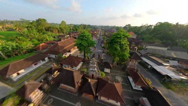 Cinematic FPV drone flight over traditional village in Bali, Indonesia, starting above local temple and continuing along decorated street surrounded by lush tropical greenery and village houses.