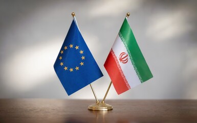 European Union and Iran national flags standing proudly side by side on a small golden base on a polished wooden desk against a plain light gray background with soft studio lighting creating a formal 