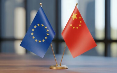 Two small desk flags representing the European Union and China stand proudly side by side on a polished wooden table in a brightly lit modern office environment with a blurred background suggesting im