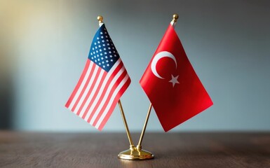 Miniature flags of the United States of America and Turkey stand proudly side by side on a dark wooden table A softly lit light grey background adds depth and a professional mood Gentle ambient light 