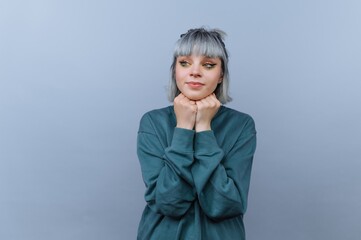 Young woman with gray hair and bangs in a teal sweater, posing thoughtfully against a light blue...