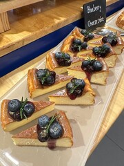 Close up of sliced blueberry cheesecake arranged in a row with fresh blueberries and sauce drizzle, showing creamy texture and warm bakery display atmosphere.