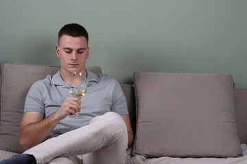 portrait of a young white male sitting on a couch and looking at a glass of white wine, drinking white wine, copy space background