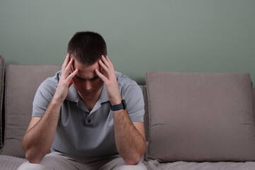 stressed young man having headache, white male sitting on couch and holding his head, thinking about problems