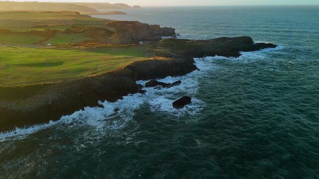Aerial drone flying forward over the ocean toward a coastal cliff at sunset with waves crashing against the coast in Spain North Coast