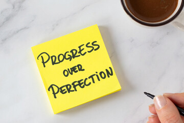Progress over perfection, handwritten inspirational quote on a sticky note with coffee cup. Top view. The concept of personal growth, success, goals, and action.