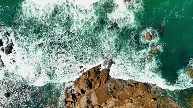 Aerial drone birds-eye view flying right over a rocky coast with ocean waves and white sea foam in Ancora Portugal