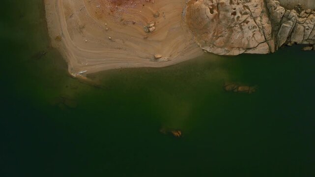 Aerial drone birds-eye view flying left and curving over a greenish lake with rocky and sandy shoreline in Spain