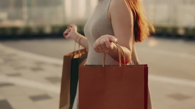 Crop view of woman with shopping bags hanging on arms walks across urban square slow motion. Young female carries purchases in paper packages at holiday sale in town district