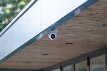 a white dome security camera mounted on the wooden-paneled underside of a modern building's roof overhang, with a blurred green background.
