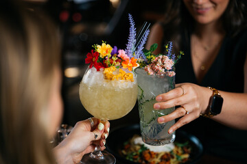 Two women are holding up their drinks, one of which has a flower garnish