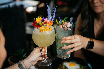 Two women are holding up glasses of drinks, one of which has a flower garnish