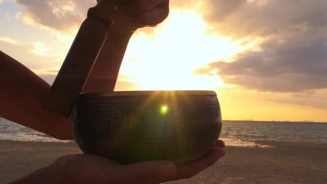 Woman's hands playing a traditional tibetan singing bowl with a mallet on a tranquil beach, creating soothing sounds for meditation and relaxation against a beautiful golden sunset over the sea