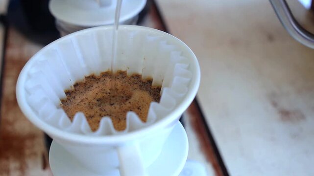 Barista carefully pouring hot water from a gooseneck kettle into a ceramic dripper with a paper filter, starting the blooming process for a delicious morning specialty coffee