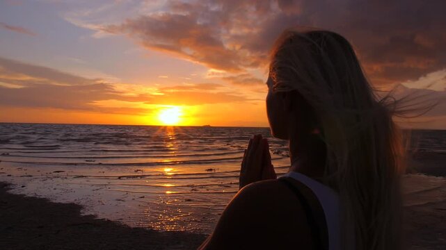 Silhouette of a spiritual woman practicing yoga and meditation on the beach, placing her hands in a namaste prayer pose against the beautiful golden sunset reflecting on the ocean water