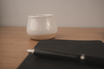 A black notebook lies open with a pen resting on top, accompanied by a white coffee cup filled with dark coffee. This setup invites creativity and reflection