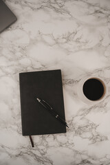 A black notebook lies open beside a sleek pen on a smooth marble surface. Next to it sits a white cup filled with dark coffee, inviting moments of inspiration and creativity