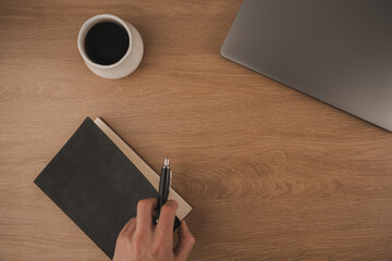 A hand writes notes in a notebook at a home workspace. A cup of coffee sits nearby, next to a laptop on a wooden surface, creating a focused atmosphere for productivity