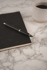 A black notebook lies open with a pen resting on top, accompanied by a white coffee cup filled with dark coffee. This setup invites creativity and reflection