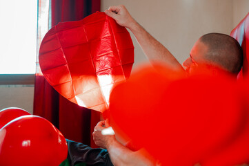 Man inflating heart-shaped balloons in a bedroom on a red bed, preparing a romantic setting for Valentine’s Day.	
