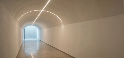 A long, minimalist white tunnel with curved walls and illuminated ceiling lights leading to a bright opening at the end.