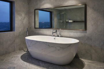 A modern freestanding bathtub with white marble pattern in a gray concrete bathroom with large mirror and window view.