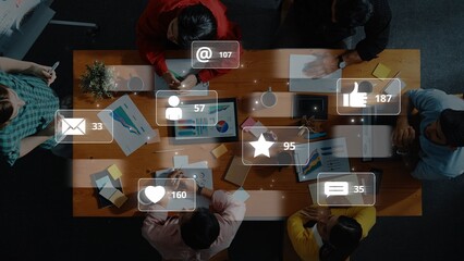 An overhead view captures a diverse group of professionals collaborating at a table, surrounded by digital interaction icons displaying various engagement metrics. Raster