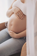 A woman sits in a chair during daylight, gently resting her hands on her round belly. Natural light comes through a window, creating a warm atmosphere in the room