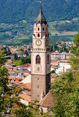 Panoramic view of Merano, Italy, with historic church tower, red rooftops, and lush alpine mountains in the background on a sunny day, showcasing European town charm.