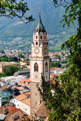 Panoramic view of Merano, Italy, with historic church tower, red rooftops, and lush alpine mountains in the background on a sunny day, showcasing European town charm.