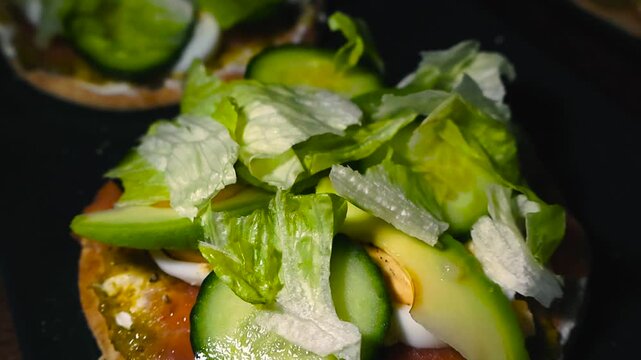 Close-up glide over colorful open-faced sandwich on dark plate topped with cucumber, lettuce, tomato and salmon with shallow depth of field and bokeh