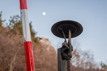 Bell detail on traditional red-and-white railway crossing barrier stands raised beside a quiet rural road. Weathered metal mechanics and chains contrast with bare autumn trees and earthy hills in the