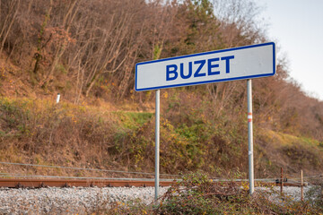 Sign board for Buzet train station at golden hour, with stone buildings and a vintage platform beside curving tracks. rural Istrian scene.