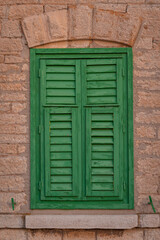Vibrant green wooden shutters with peeling paint stand closed against a textured stone wall. A heavy stone arch and sill frame the window, accented by small metal latches on either side of the base.