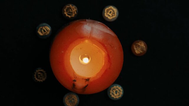 Top view of a burning orange candle surrounded by engraved chakra stones, Atmospheric spiritual video of candle flame and chakra energy symbols on black