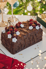 Christmas cake with cherries, cream and chocolate on holiday background with glasses, candy and xmas tree. Merry Christmas and Happy New Year food, bokeh, lights