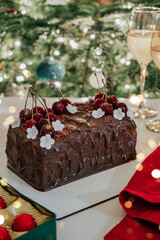 Christmas cake with cherries, cream and chocolate on holiday background with glasses, candy and xmas tree. Merry Christmas and Happy New Year food, bokeh, lights