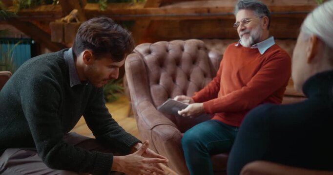 Sad and lonely young man sharing personal problems during group therapy, receiving warm supportive hug from therapist after session, showing relief and gentle smile in calm professional environment.