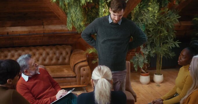 Nervous and insecure participant speaking to group during psychotherapy, rubbing his hands and avoiding eye contact, clearly uncomfortable while sharing personal difficulties in calm supportive settin