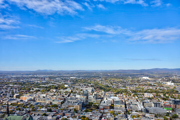 A wide-angle aerial view of Quebec city under a bright blue sky, showing dense urban development, residential and commercial zones, highways, and distant mountains on the horizon.