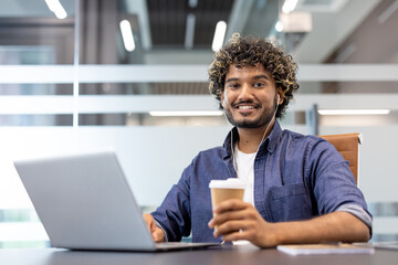 Portrait of a young Indian man sitting at a desk in the office wearing headphones, holding a cup of coffee in his hand and looking at the camera with a smile