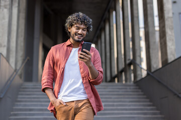 Indian young man in red shirt standing near building, holding hand in pocket and using mobile phone