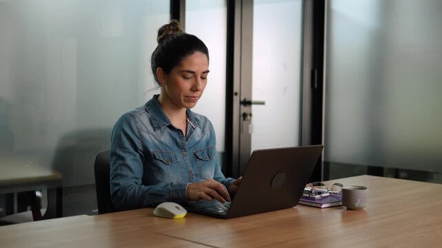 Young professional woman in denim shirt smiling with excitement while reading good news on laptop at desk in modern office