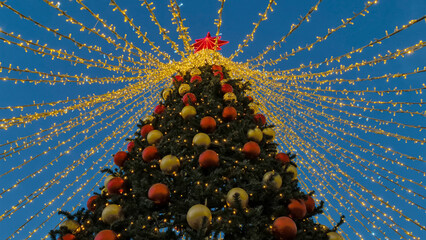 A festive Christmas tree decorated with colorful balls and a red star in the town square of Zelenogradsk, bright New Year's lights garland against the backdrop of a twilight blue sky.