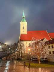 Illuminated St. Martin's Cathedral tower rising above a glowing tree in Bratislava, Slovakia, casting reflections on wet cobblestones during a foggy Christmas night in the historic city center.