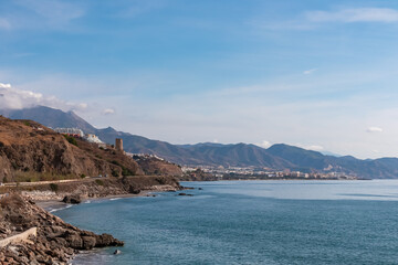 Scenic view of the Torrox Costa coastline featuring a winding road along rocky cliffs and historic Torre de Calaceite, overlooking the calm blue Mediterranean Sea and mountains in Andalusia, Spain.